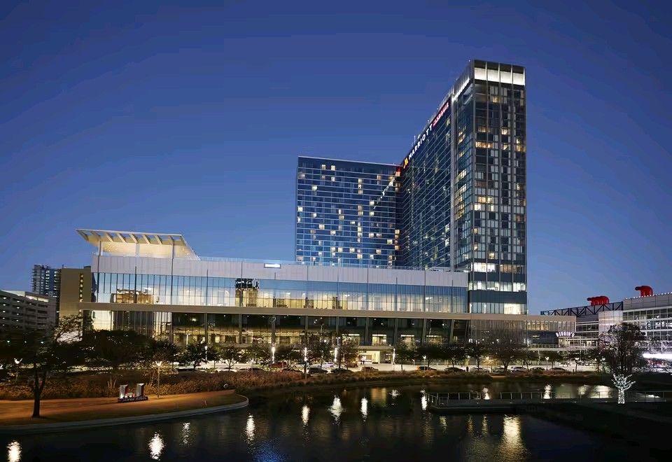 Modern, illuminated high-rise building with glass facade reflected on a calm water body at night.